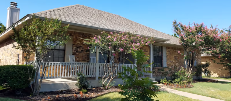 Front exterior of a brick senior living facility with a wrap porch and flowering trees.