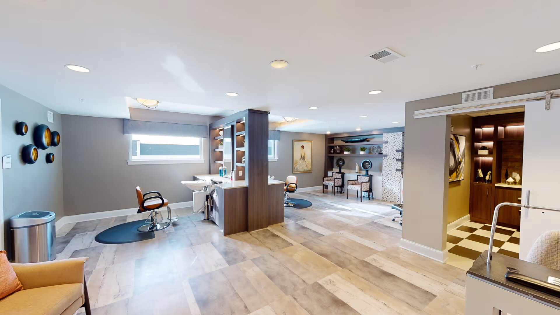 A spacious, well-lit interior room with two salon-style chairs positioned in front of mirrors and sinks, a seating area with two patterned chairs and a small table, decorative wall art, and a partially open sliding door revealing a room with checkered flooring and display shelves.