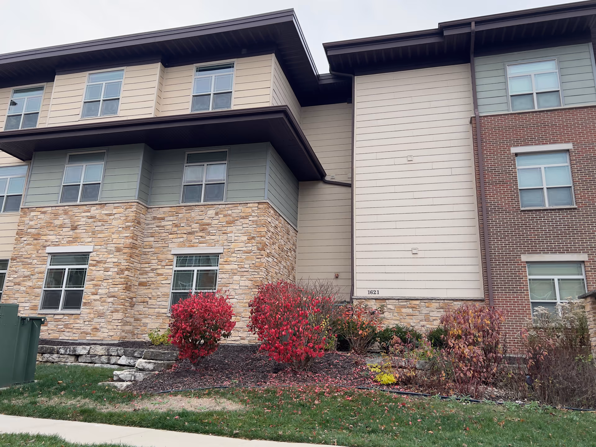 Exterior view of a multi-story senior living facility building with a combination of stone, brick, and siding on the facade. There are several windows and landscaping with red and green bushes in front of the building. The address number 1621 is visible on the siding.