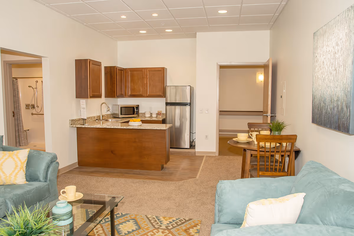 Interior view of a senior living facility apartment showing a small kitchen with wooden cabinets, a stainless steel refrigerator, microwave, and granite countertop. Adjacent to the kitchen is a living area with teal armchairs, a glass coffee table, and a patterned rug. A small round dining table with two wooden chairs is also visible. There is a bathroom with a shower visible through an open door on the left side.