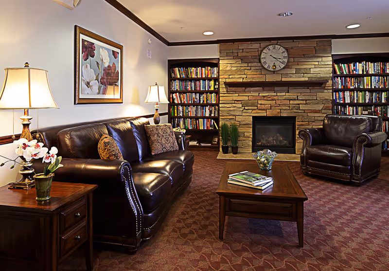 A cozy living room with a dark brown leather sofa and armchair, a wooden coffee table with books and a decorative bowl, two table lamps on wooden side tables, a framed floral painting on the wall, a stone fireplace with a clock above it, and bookshelves filled with books on either side of the fireplace.