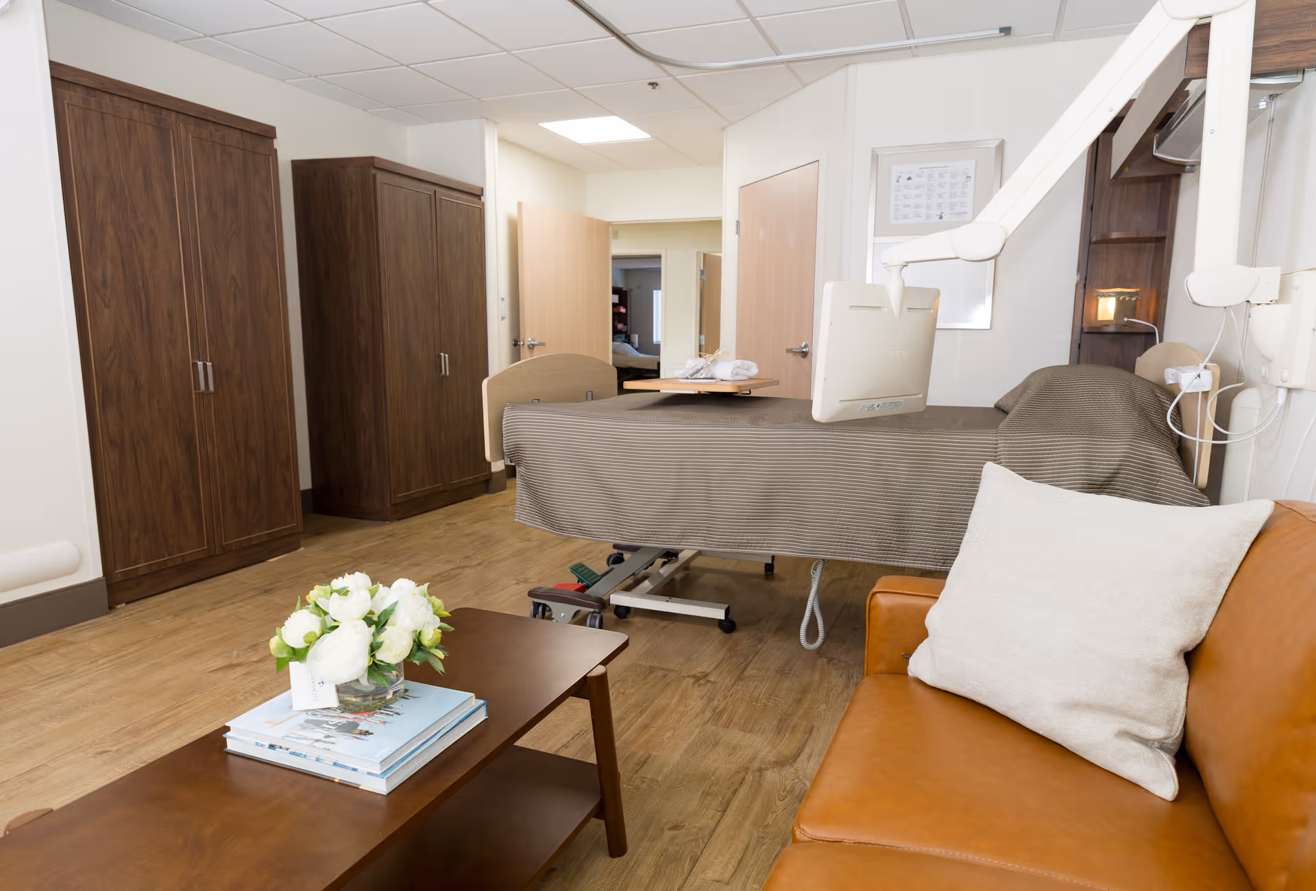 A senior living facility room featuring a hospital-style bed with a brown striped blanket, a mounted monitor, and a brown leather couch with a white pillow. There are two wooden wardrobes against the wall, a wooden coffee table with a vase of white flowers and books, and an open door leading to another room with a bed visible.