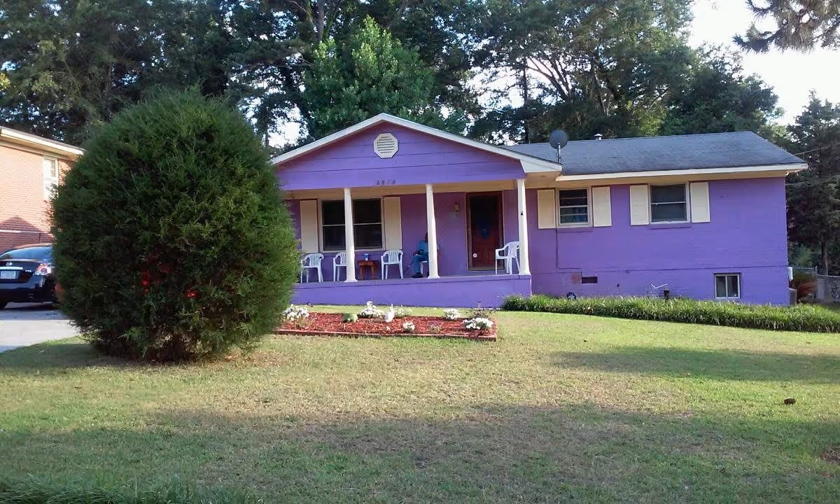 Front exterior view of a single-story purple house with a small porch featuring white chairs. There is a large bush in the front yard and a flower bed with white flowers. Trees and a parked car are visible in the background.