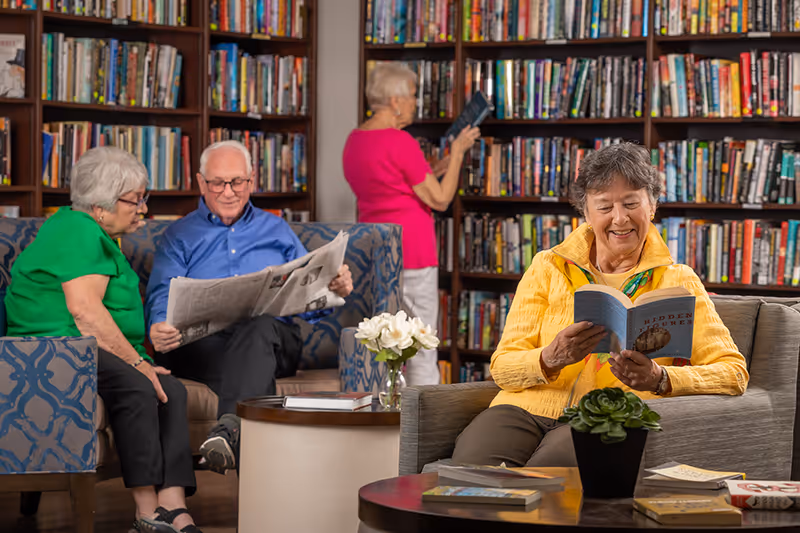 Four elderly individuals in a library setting. One woman in a yellow jacket is sitting on a couch reading a book, a man in a blue shirt and a woman in a green shirt are seated together on a patterned couch with the man reading a newspaper, and another woman in a pink shirt is standing and selecting a book from the bookshelf behind them.