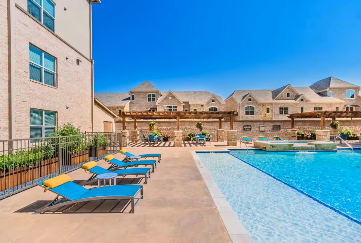 Outdoor swimming pool area with blue lounge chairs, pergolas, and a brick residential building under a clear blue sky.