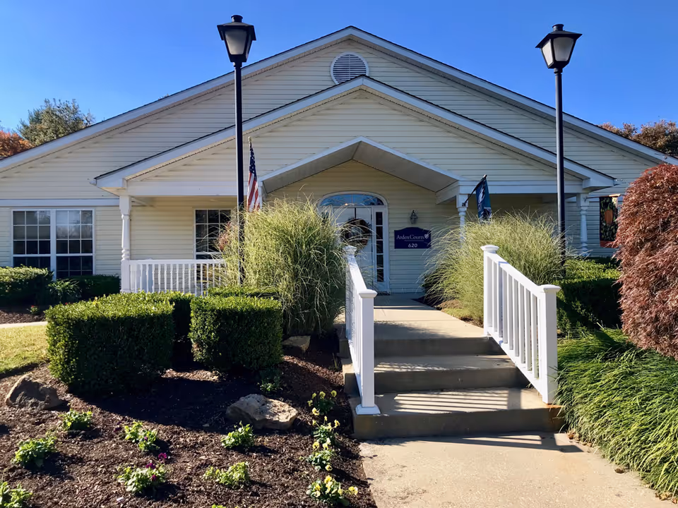Front exterior view of a single-story building with beige siding, a small porch with white railings, two black lamp posts, and landscaped bushes and flowers along the walkway leading to the entrance. An American flag is displayed near the porch, and a sign next to the door reads Arden Courts 620.