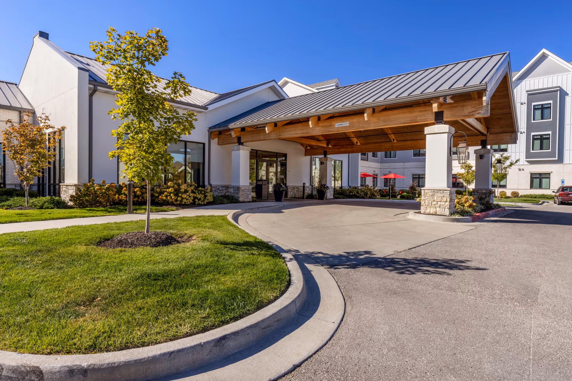 Exterior view of Meadowbrook Senior Living facility entrance with a covered drop-off area supported by stone and wooden pillars, surrounded by green grass, small trees, and shrubs under a clear blue sky.