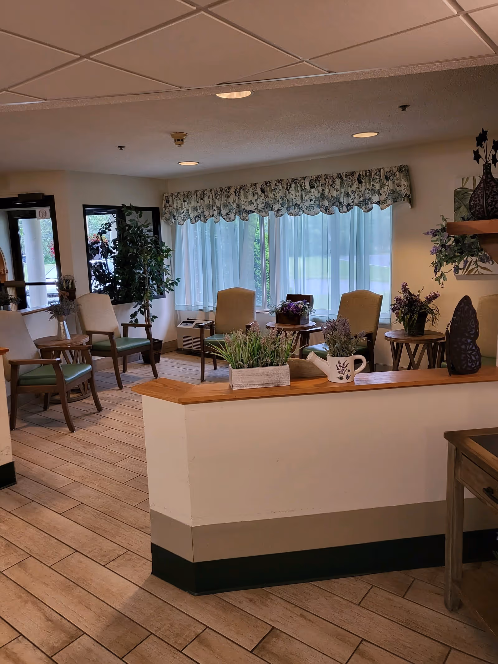 A cozy sitting area in a senior living facility with several cushioned chairs arranged around small wooden tables. The room has large windows with sheer curtains and floral valances, allowing natural light to brighten the space. Decorative plants and floral arrangements are placed on tables and shelves, creating a welcoming atmosphere. The floor is tiled with a wood-like pattern, and the ceiling has recessed lighting.