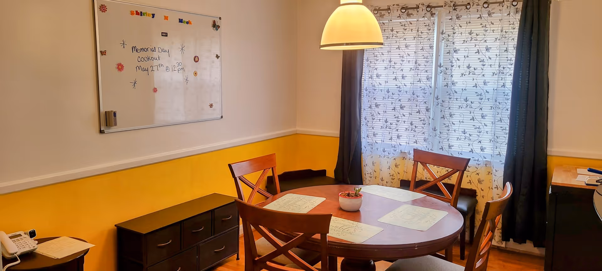 A small dining area with a round wooden table and four chairs. The table has placemats and a small potted plant in the center. A hanging light fixture is above the table. Behind the table is a window with sheer floral curtains and dark drapes. On the left wall, there is a whiteboard with colorful letters spelling 'Shirley & Mack' and a note about a Memorial Day cookout. Below the whiteboard is a black cabinet and a small table with a telephone and papers.