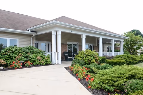 Exterior view of a single-story building with a covered porch supported by white columns. The porch has seating with black chairs. The building is surrounded by well-maintained landscaping including green shrubs and flowering plants. The sky is clear and the overall scene is bright and inviting.