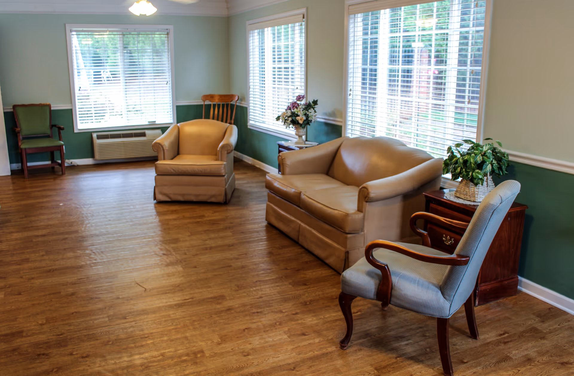 A living room with wooden flooring, featuring a beige leather sofa, a beige armchair, a wooden chair, and a blue upholstered chair. There are two side tables with decorative plants and flowers, and large windows with white blinds letting in natural light.