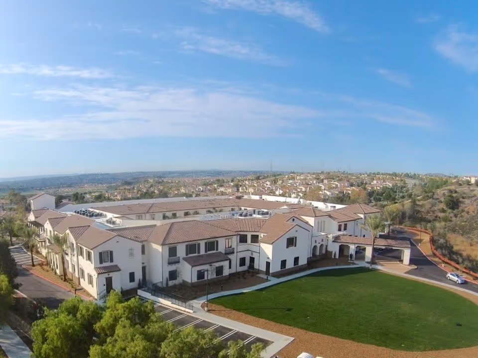 Aerial view of Westmont at San Miguel Ranch, a large senior living facility with white buildings and brown tiled roofs, surrounded by green lawns and trees under a blue sky.