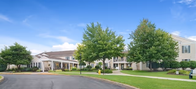 Exterior view of Brookdale Summerfield senior living facility showing a large, light-colored building with multiple windows, surrounded by green trees and well-maintained lawns under a blue sky with some clouds.