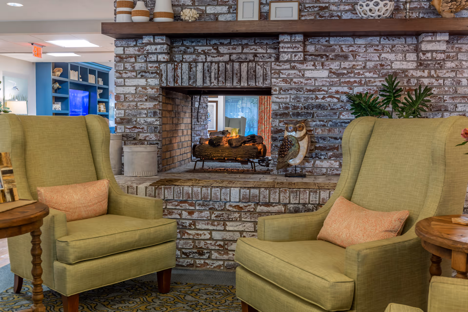 Two green upholstered armchairs face a double-sided whitewashed brick fireplace in a cozy senior living lounge.