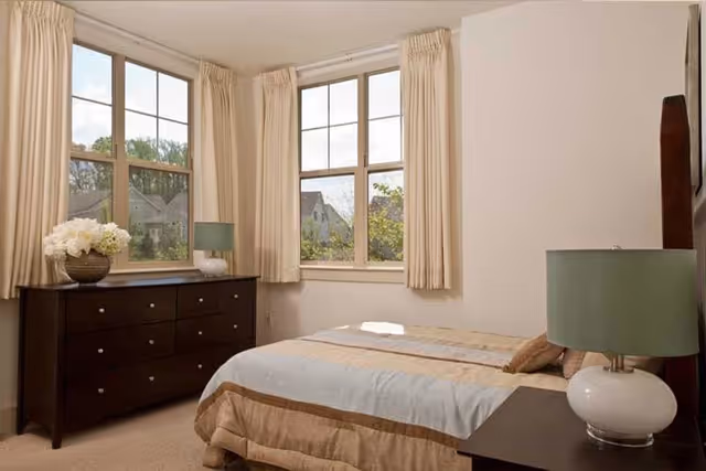 A bedroom with a bed covered in beige and light blue bedding, a dark wooden dresser with a vase of white flowers, two windows with beige curtains, and a nightstand with a green lamp.