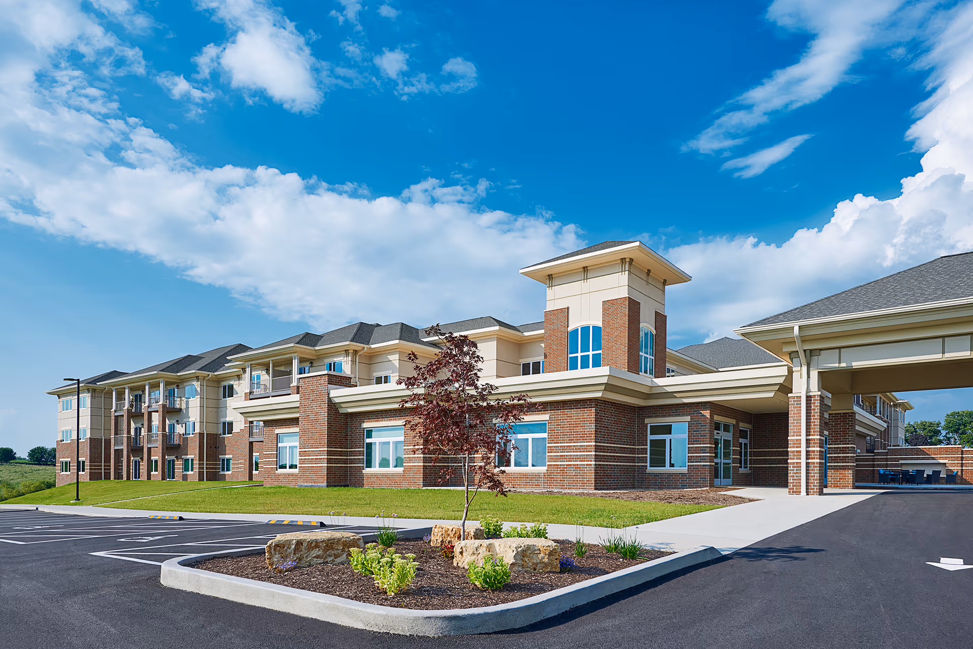 Front exterior of a multi-story brick senior living facility with a covered drop-off, landscaping, and parking lot under a blue sky.