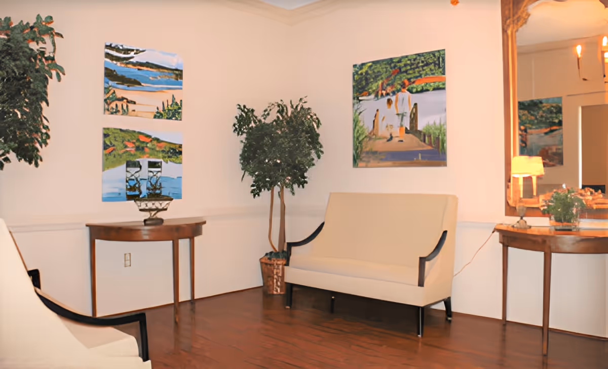 A cozy sitting area with two beige upholstered benches with dark wooden arms and legs, a wooden side table with a lamp and a small plant, a large wall mirror with a wooden frame, two potted plants, and three colorful paintings on the walls depicting outdoor scenes.