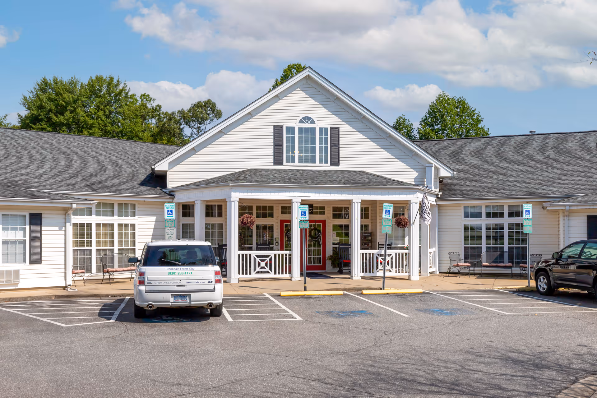 Front exterior view of Brookdale Forest City facility showing a white building with a peaked roof, large windows, a covered entrance with columns, and a parking lot with handicap parking spaces and vehicles.