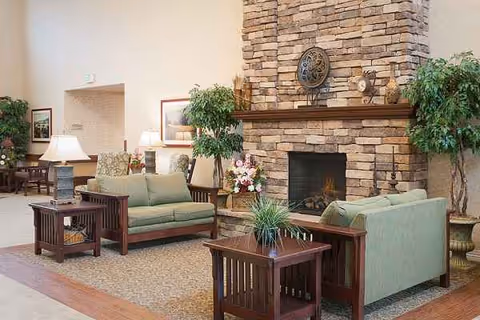 A welcoming lounge with green couches, wooden tables, plants, and a stone fireplace.