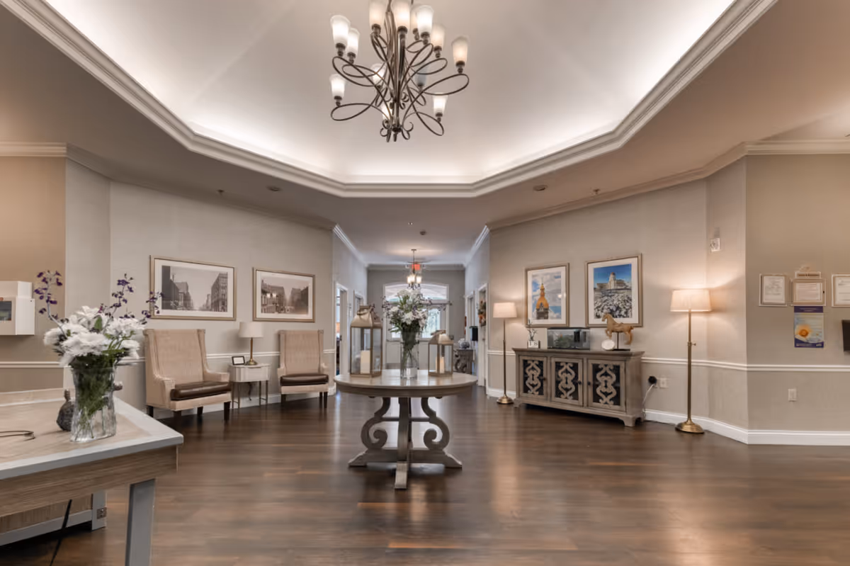 A spacious and elegantly decorated common area with wooden flooring, beige walls, and a tray ceiling with a chandelier. The room features a round wooden table with a flower arrangement in the center, two beige upholstered chairs with a small side table between them, framed pictures on the walls, a decorative sideboard with lamps and ornaments, and floor lamps providing warm lighting.