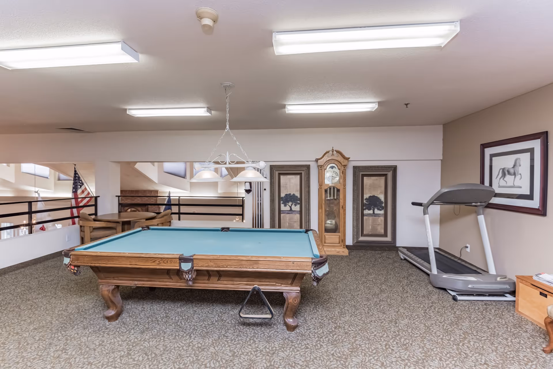 A recreational room with a pool table in the center, a treadmill on the right, a grandfather clock and two framed pictures on the back wall, and a small round table with chairs near a railing overlooking a lower level with flags.