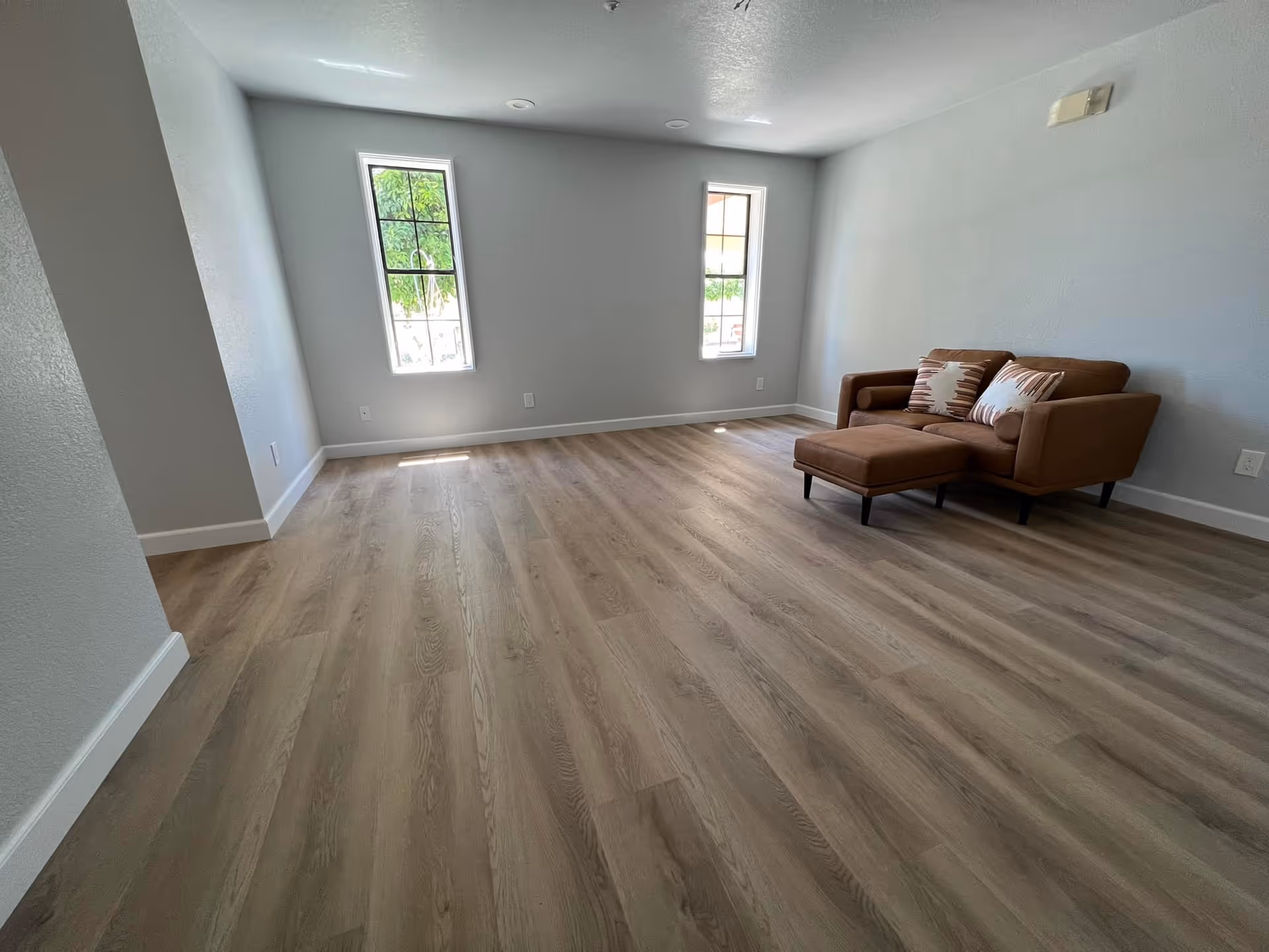 A bright and spacious room with light wood flooring, two tall narrow windows letting in natural light, and a brown upholstered armchair with matching ottoman and decorative pillows against a light gray wall.