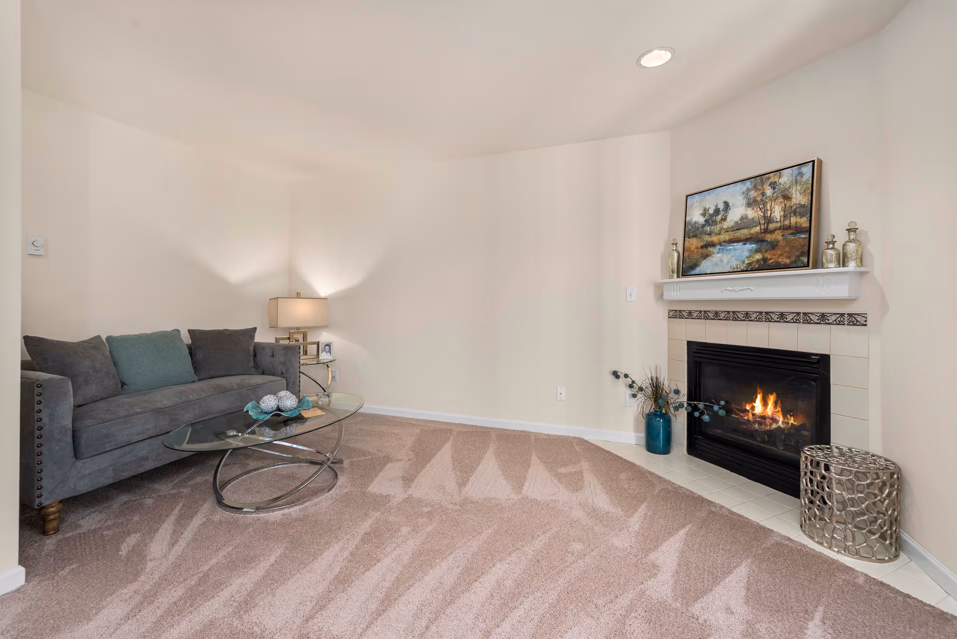 Cozy living room with a gray sofa, glass coffee table, and a lit fireplace under a painting.