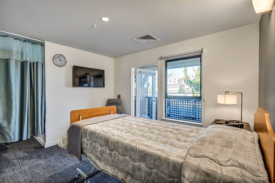 A bedroom in a senior living facility with a single bed covered in a beige patterned blanket. The room has a wall-mounted TV, a clock, a window with a view outside, a small bedside table with a lamp, and a chair in the corner. There is a curtain partially covering an area on the left side of the room.