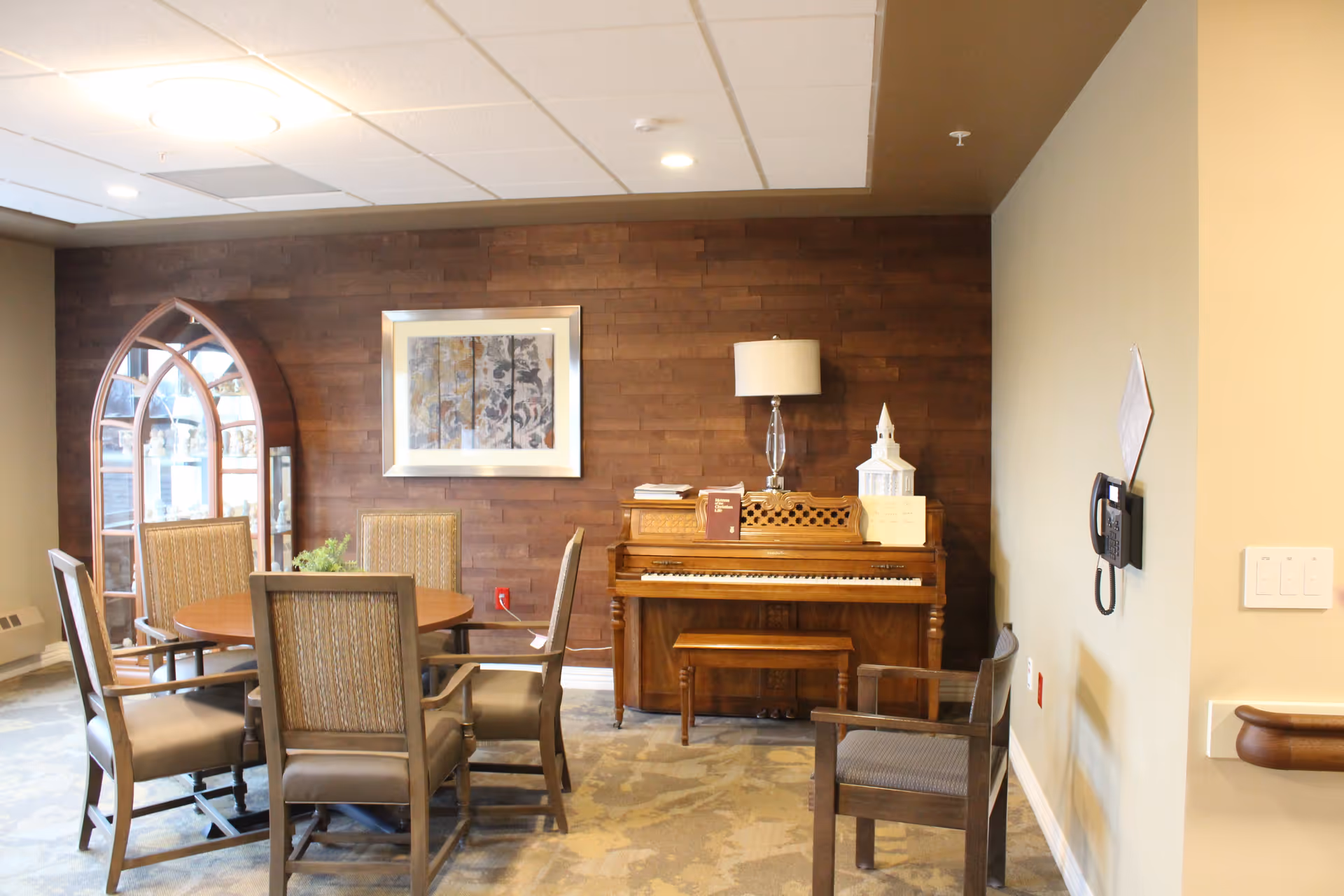 A cozy interior room with a round wooden table surrounded by five upholstered chairs. Against a wooden paneled wall is an upright piano with a bench, a table lamp, and decorative items on top. A framed artwork hangs above the piano, and a wooden display cabinet with glass doors is visible in the corner. The room has a carpeted floor and a ceiling with recessed lighting.