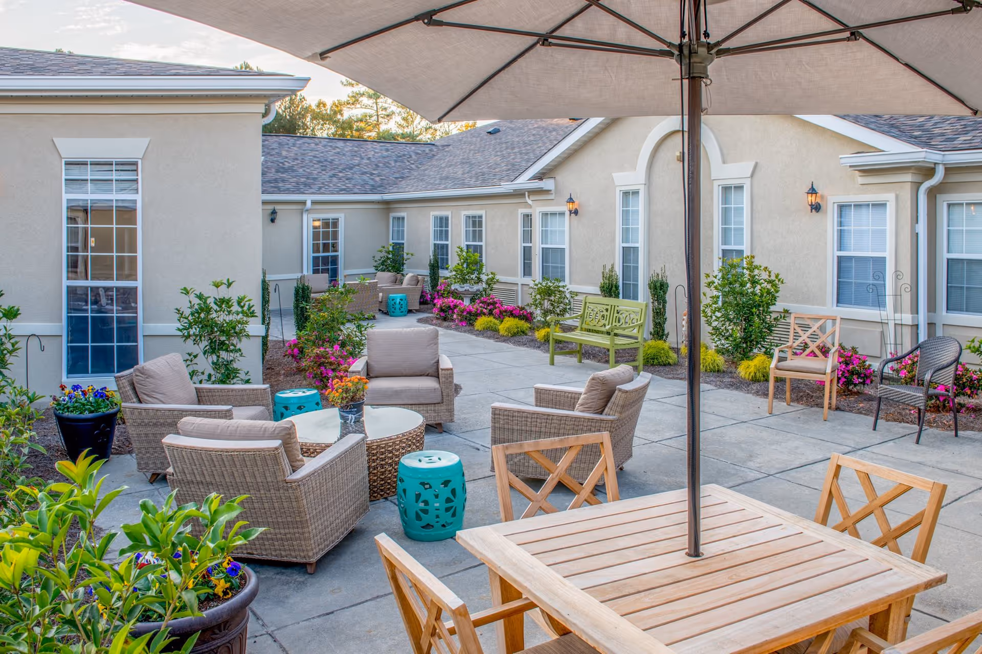 Sunny outdoor courtyard with wicker seating, a wooden dining table and umbrella, potted flowers and benches outside a beige senior living building.