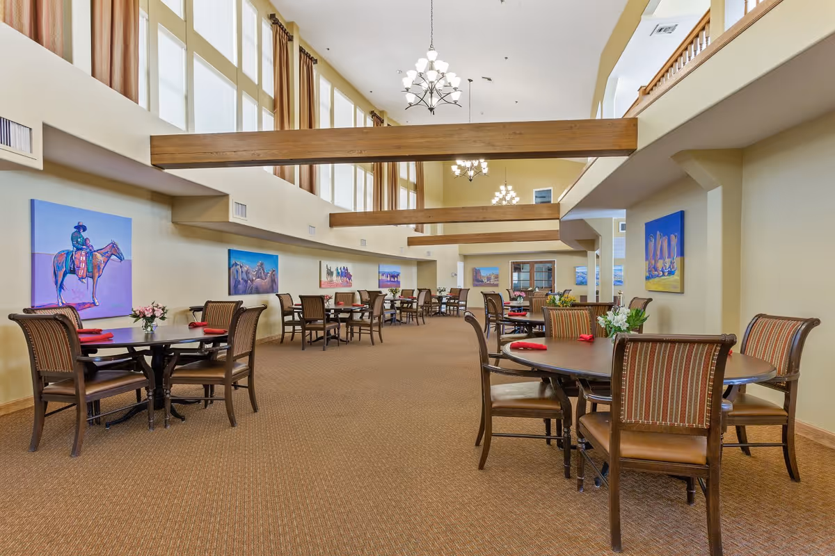 A spacious dining room with multiple round tables and chairs arranged neatly. The room features high ceilings with wooden beams and large windows near the ceiling allowing natural light to fill the space. Colorful paintings of horses and riders adorn the walls, and small flower arrangements and red napkins are placed on the tables.