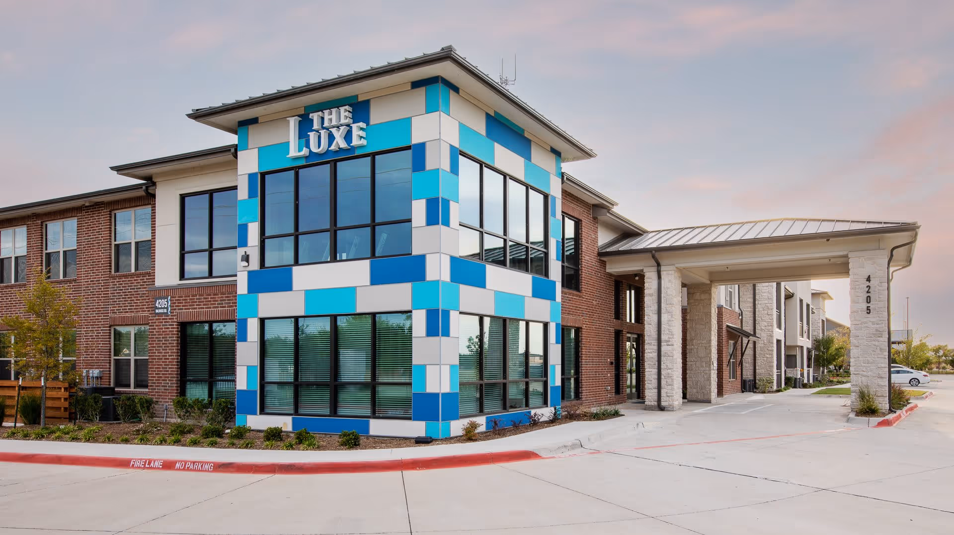 Exterior view of The Luxe at Rowlett senior living facility building with a modern design featuring blue and white paneling, large windows, and a covered entrance. The building is surrounded by a paved driveway and landscaped greenery under a partly cloudy sky.