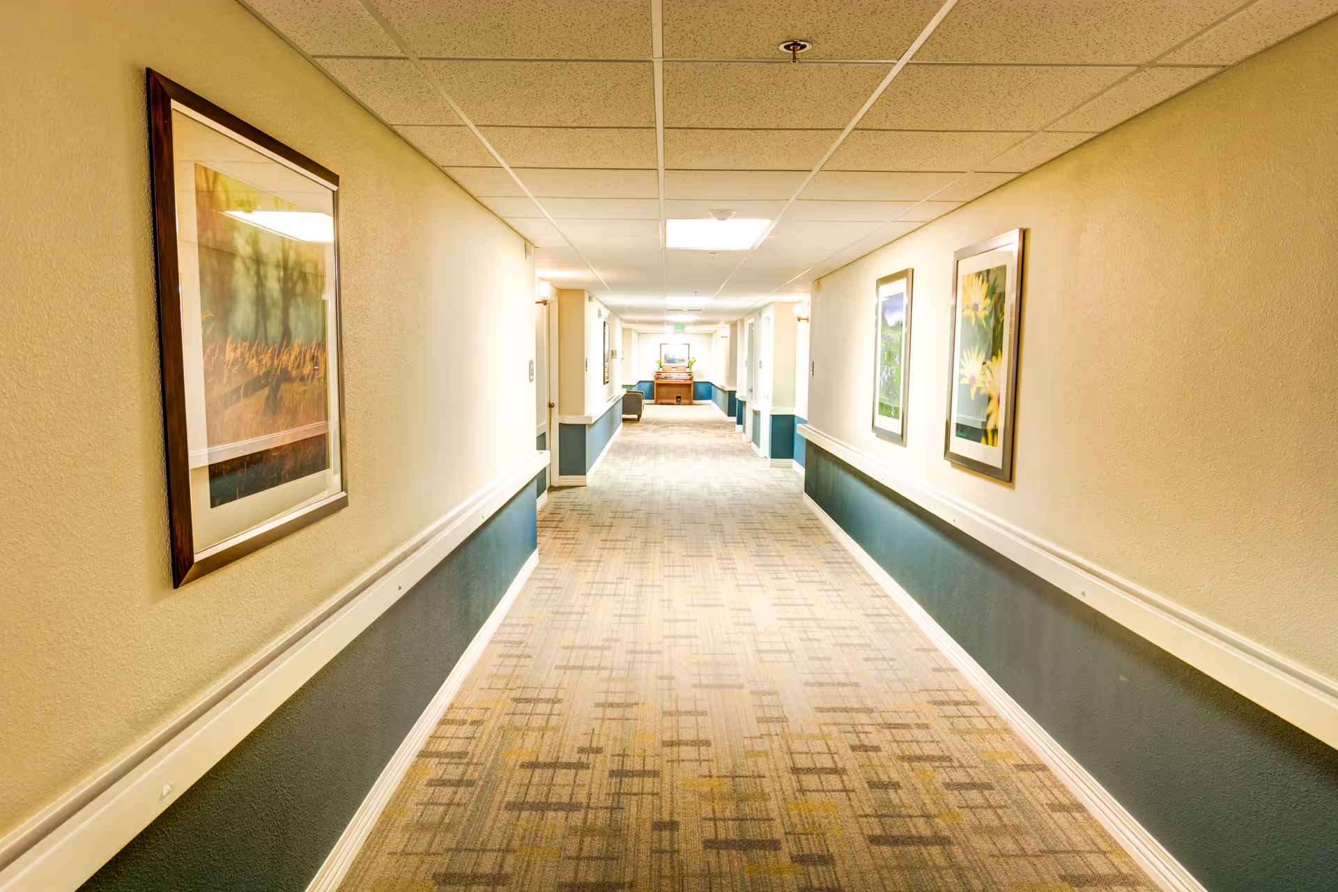 Long carpeted interior hallway in an assisted living facility lined with framed artwork and handrails.