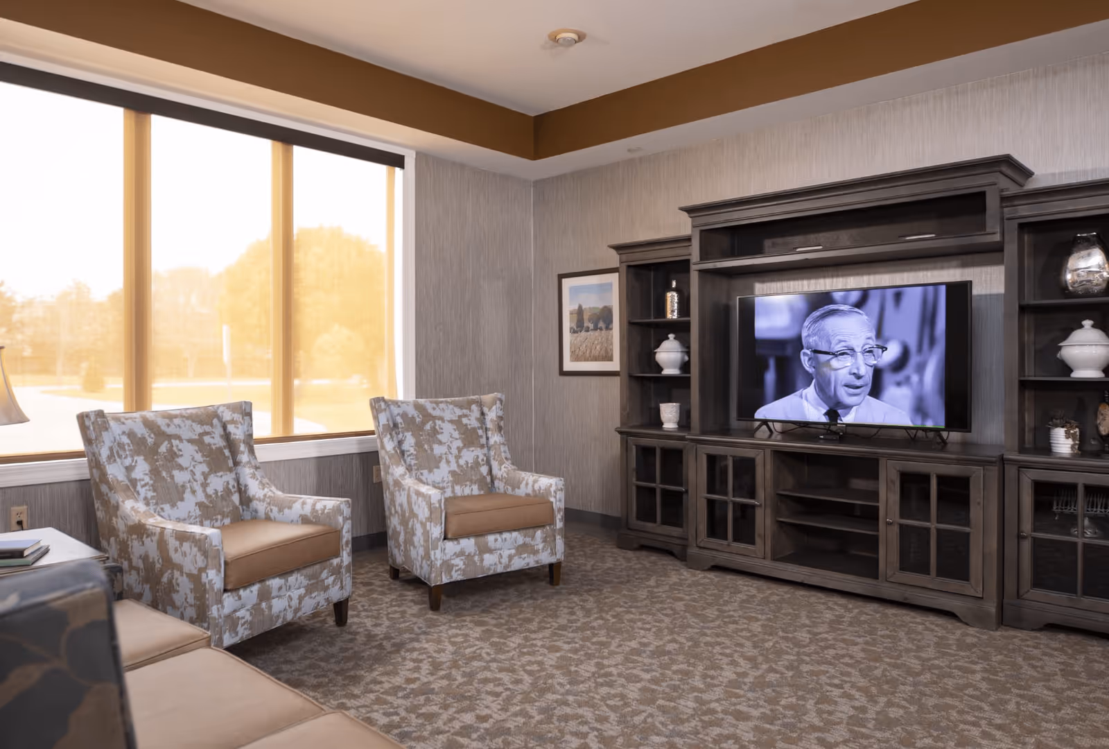 A cozy living room with two patterned armchairs and a beige sofa facing a large wooden entertainment center with a flat-screen TV displaying a black and white image of an elderly man. The room has a large window with a view of trees outside, beige carpet, and neutral-colored walls with a framed picture.
