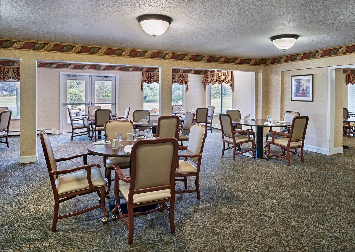 A dining room in a senior living facility with multiple round tables and cushioned wooden chairs. Each table is set with glasses, napkins, and salt and pepper shakers. The room has patterned carpet, wallpaper with a decorative border near the ceiling, and large windows with valances allowing natural light to enter. There are double glass doors leading outside and framed artwork on the walls.