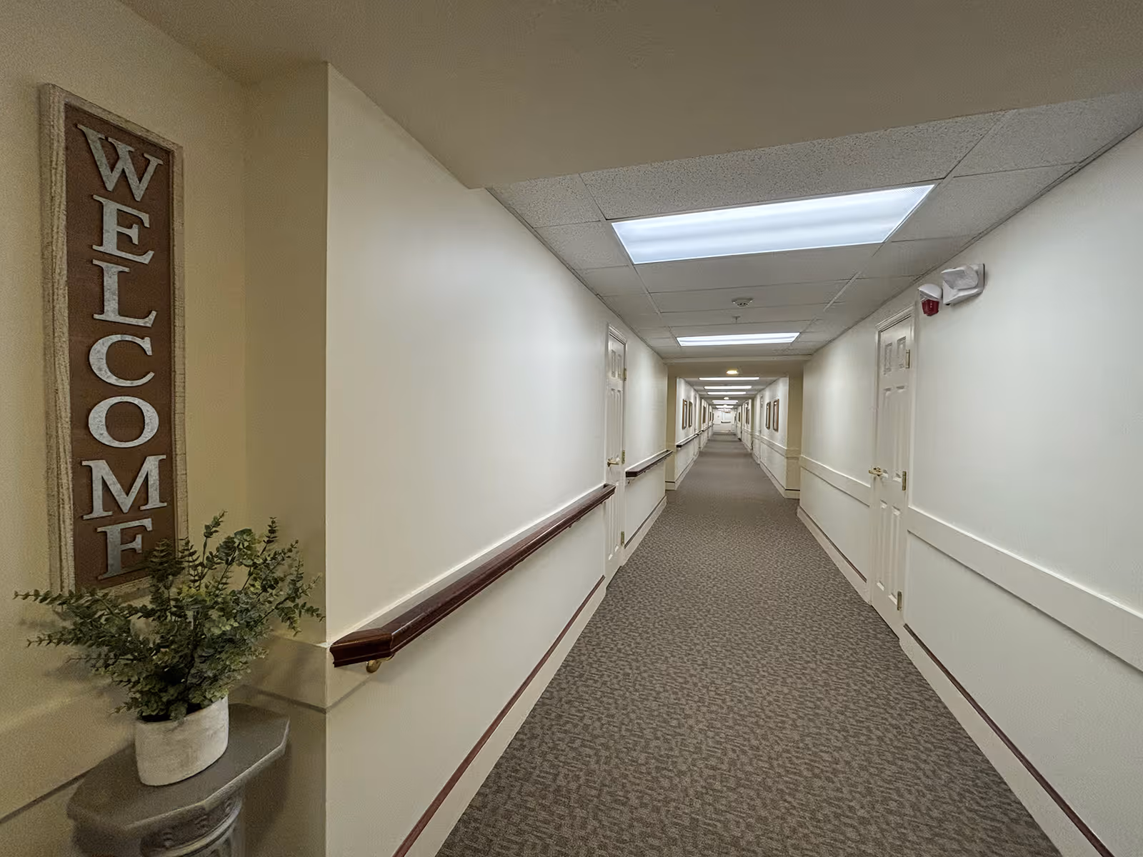Long, well-lit hallway in a senior living facility with beige walls, carpeted floor, handrails on both sides, and multiple closed doors along the corridor. A decorative 'WELCOME' sign and a potted plant are visible on the left side near the foreground.