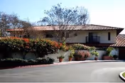Front of a low-rise stucco building with a tiled roof, driveway loop, and potted plants along the entrance.