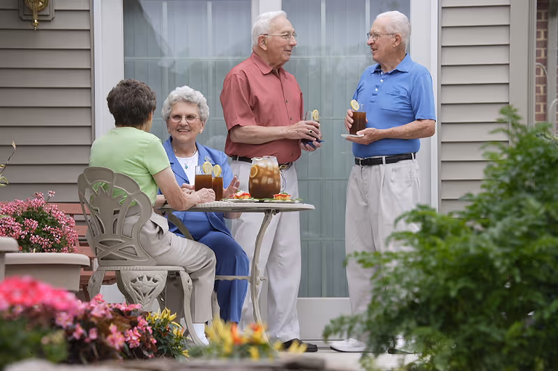 Four elderly people socializing outdoors near a table with drinks and snacks. Two women are seated, one wearing a green shirt and the other in a blue outfit, while two men stand nearby holding glasses of iced tea with lemon slices. The setting is a patio area with flowers and greenery around, in front of a building with beige siding and a glass door.