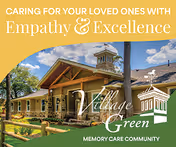 Exterior view of Village Green Memory Care Community Kingwood building with a clear blue sky and some trees in the background. The building has a wooden entrance with a peaked roof and a tower-like structure above it. Text overlay reads 'Caring for your loved ones with Empathy & Excellence' along with the Village Green logo and 'Memory Care Community'.
