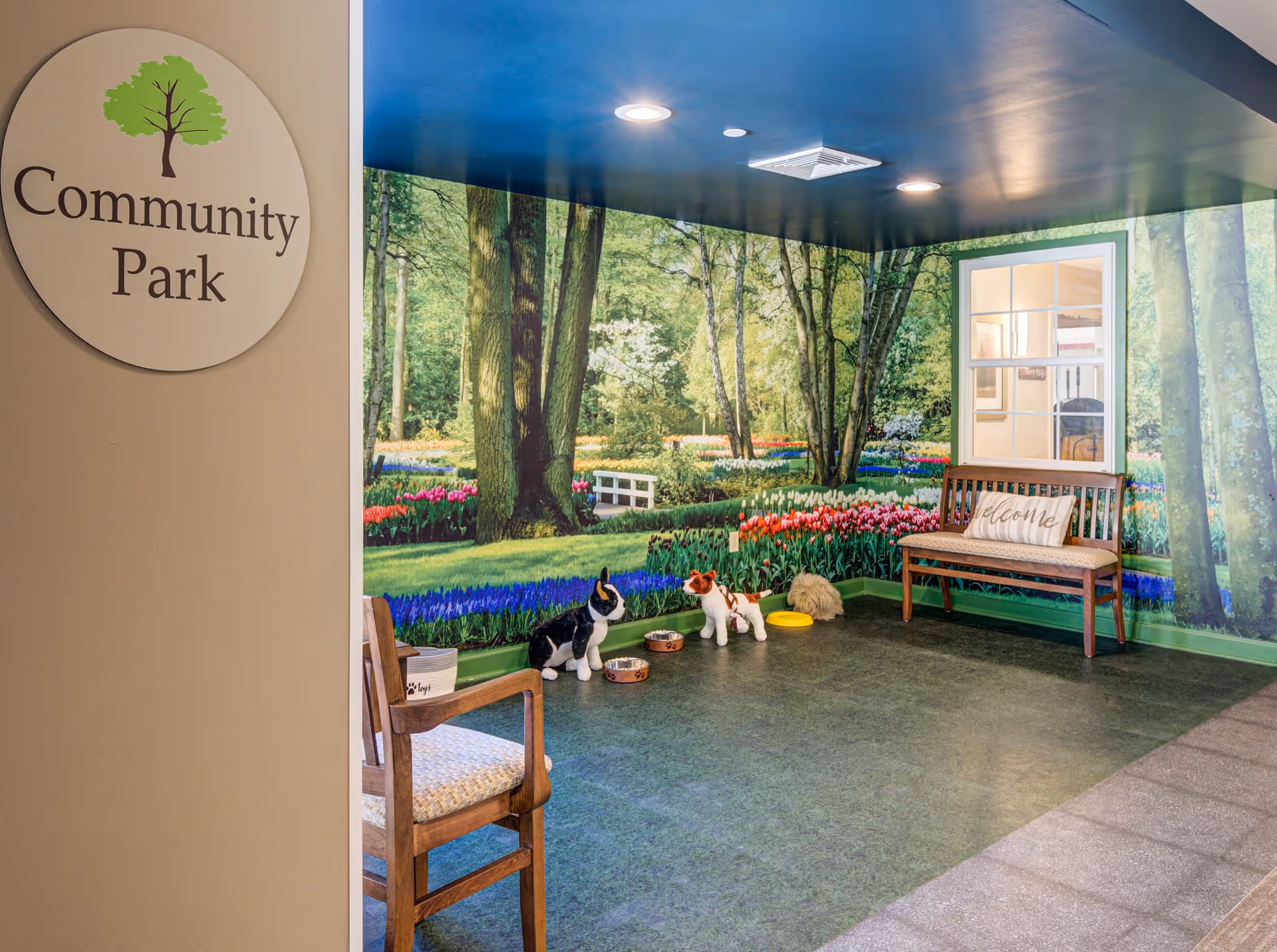 Indoor room with a mural of a park scene featuring trees and colorful flowers on the walls. The room has a wooden bench with a 'welcome' pillow, a wooden chair, and several pet bowls on the floor with stuffed animal dogs. A round sign on the wall reads 'Community Park'.