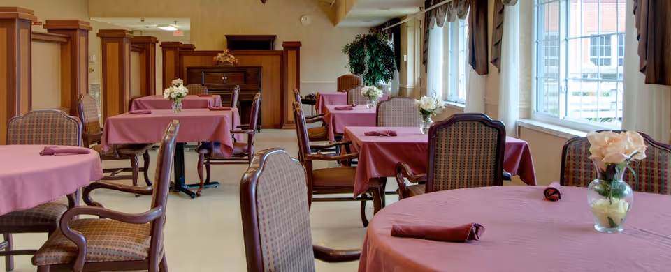 Dining room with multiple tables covered in mauve tablecloths, each set with a small vase of white flowers and folded napkins. The room has large windows with curtains letting in natural light, wooden chairs with patterned upholstery, and a piano against the far wall.