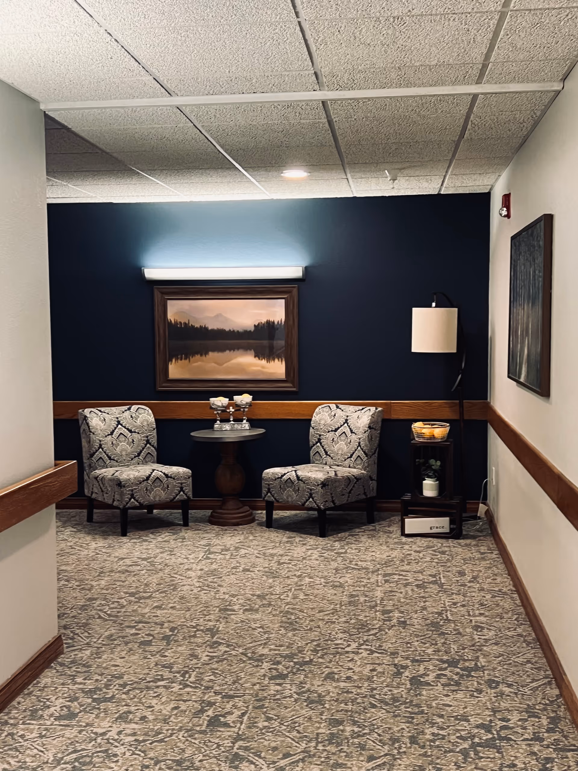 Two patterned chairs flank a small round table beneath a framed landscape painting in a hallway seating nook.
