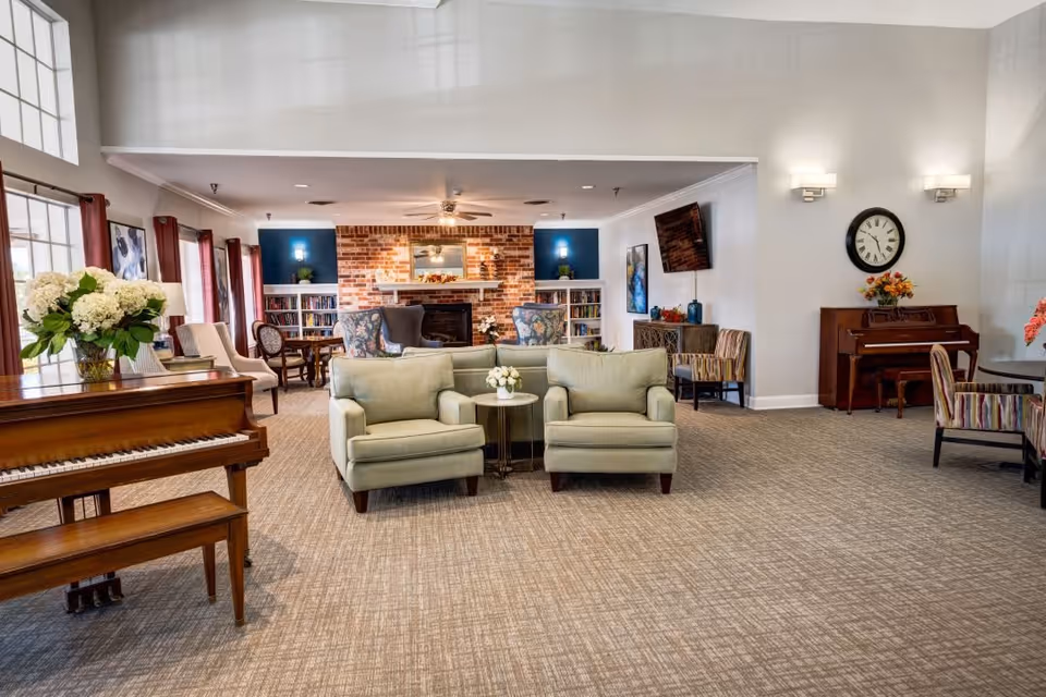 A spacious and well-lit living room area in a senior living facility featuring two light green armchairs with a small round table between them, a wooden piano with a vase of white flowers on top, a brick fireplace with built-in bookshelves on either side, a wall-mounted TV, a clock on the wall, and several chairs and tables arranged around the room.