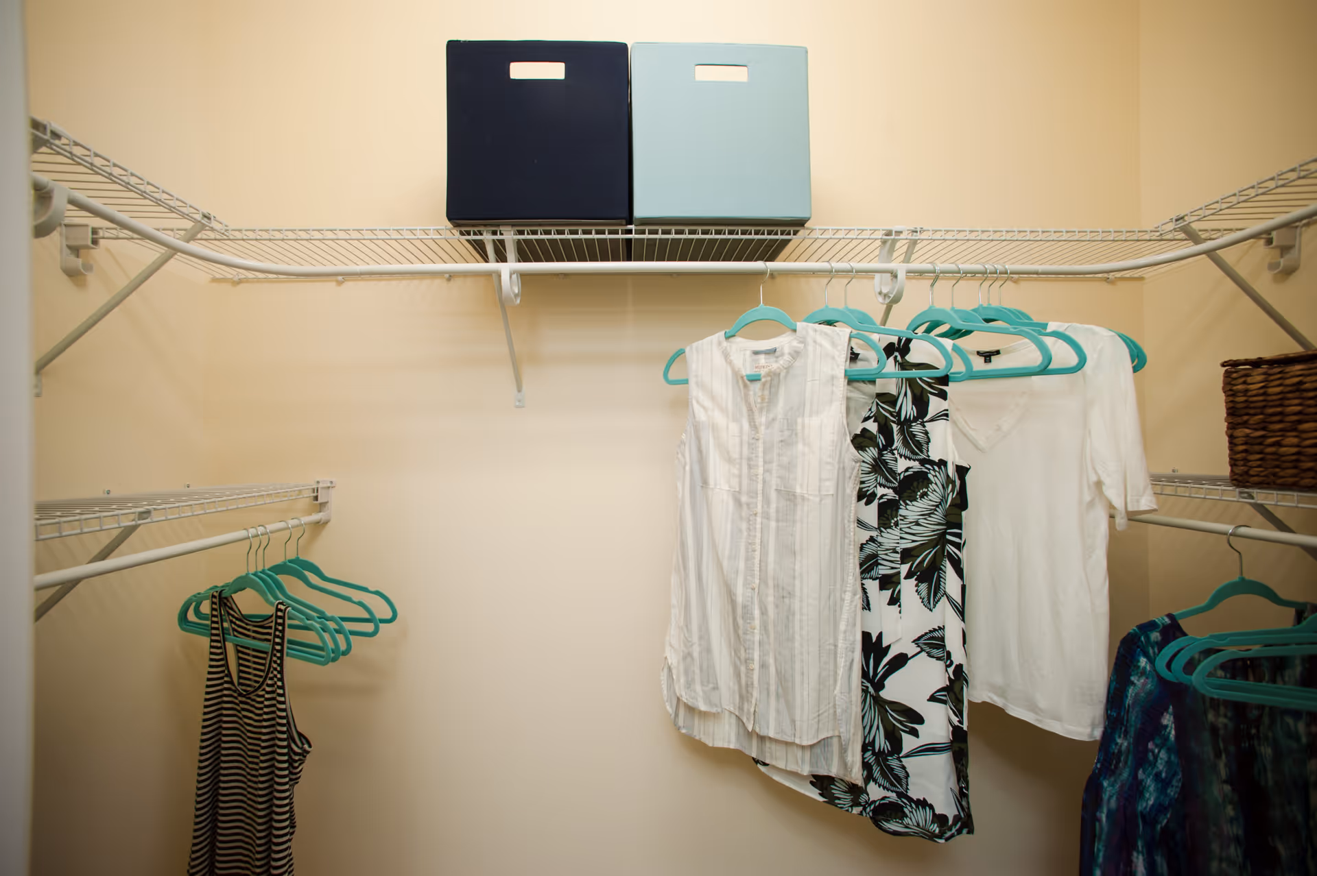Interior view of a closet with wire shelving. Several clothes hang on turquoise hangers, including a striped tank top, a white sleeveless blouse, a black and white floral sleeveless blouse, a white short-sleeve top, and a blue patterned garment. Two storage boxes, one dark blue and one light blue, are placed on the top shelf. A woven basket is on the right side shelf.