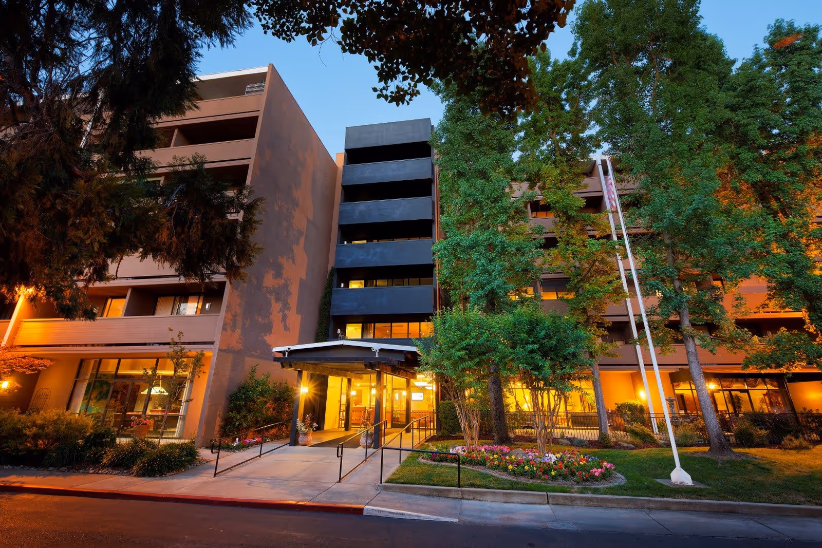Exterior view of Eskaton Land Park building at dusk with illuminated entrance, surrounded by trees and landscaped flower beds.