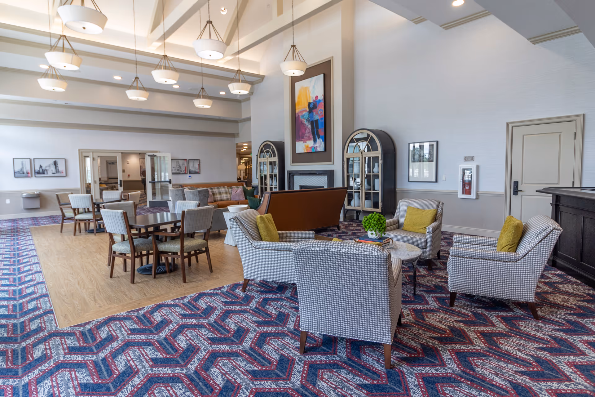 Spacious common lounge in a senior living facility with armchairs, small tables, dining tables, patterned carpet and high pendant lighting.