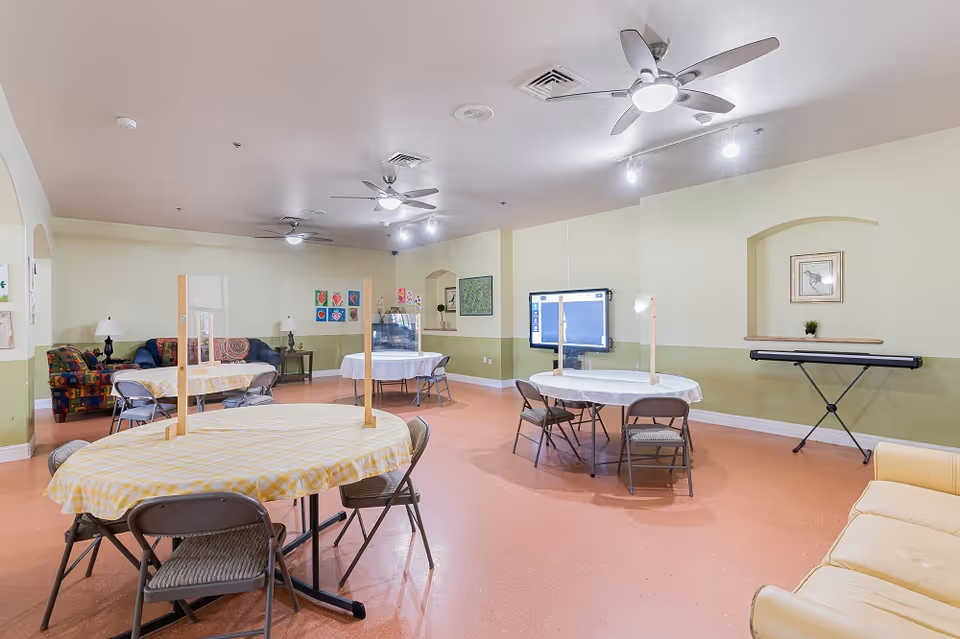 A large multipurpose common room with round tables and folding chairs, sofas, a keyboard, and a wall-mounted TV.