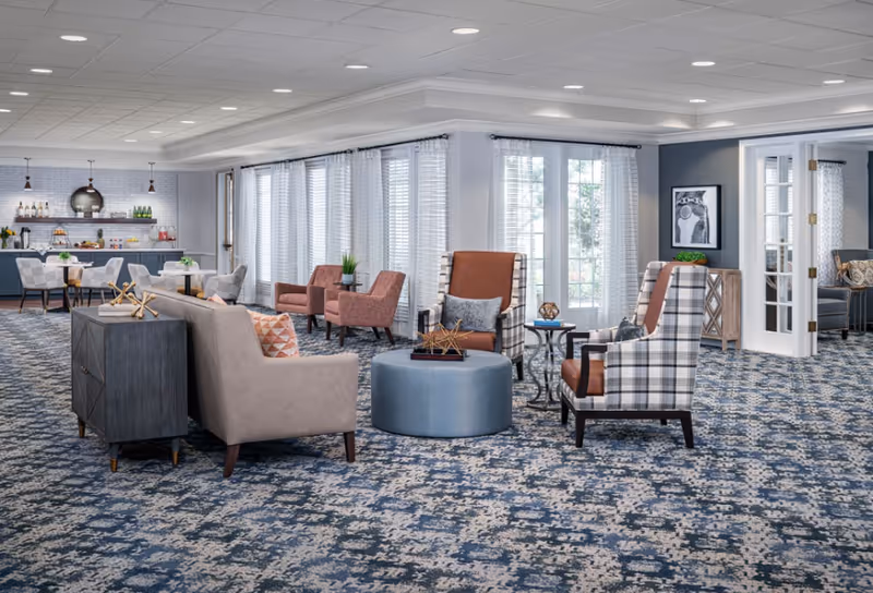 A spacious and well-lit common area with patterned blue carpet, multiple seating options including beige sofas, plaid and brown armchairs, and small tables. Large windows with sheer white curtains allow natural light to fill the room. In the background, there is a dining area with tables and chairs, and a counter with beverages and decor items.