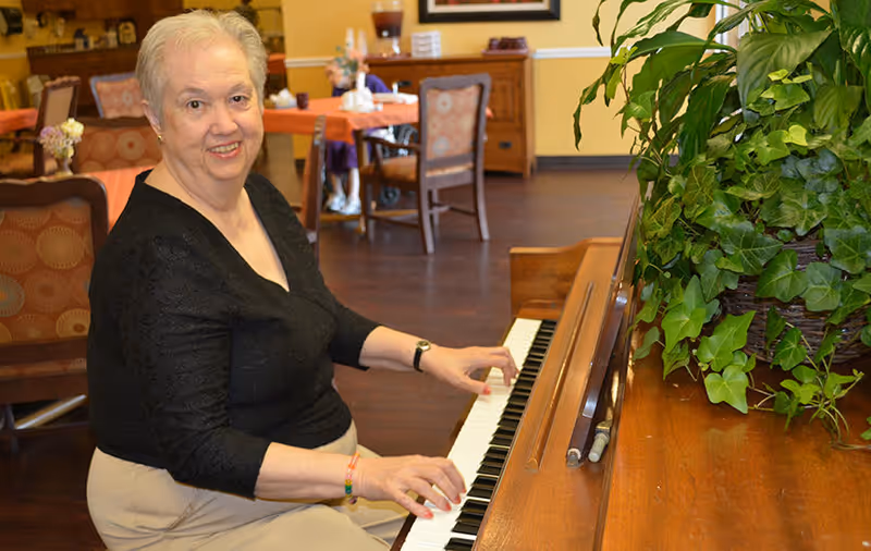An elderly woman with short white hair wearing a black top and beige pants is sitting at a wooden piano, playing it and smiling at the camera. The room has wooden floors, several tables and chairs, and a large green leafy plant on the piano.