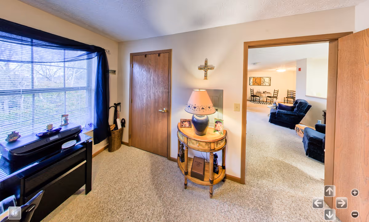 Interior view of a senior-living apartment entry area with a lamp table, closed door, window and a connected living room with armchairs.